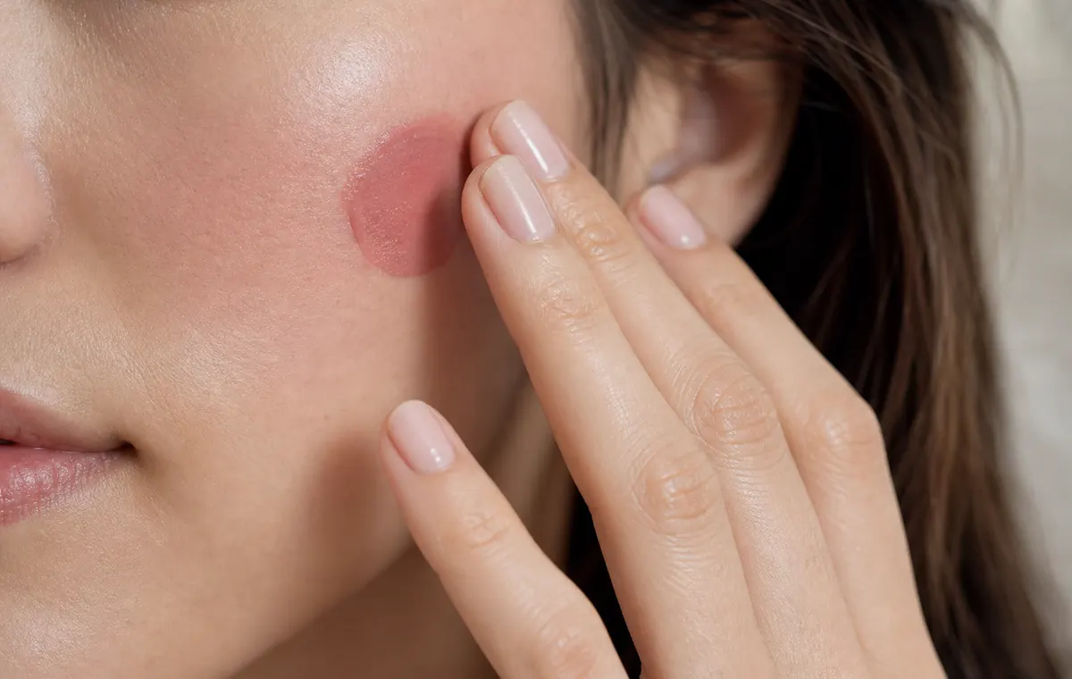 A hand tapping cream blush high on the cheek in soft daylight.